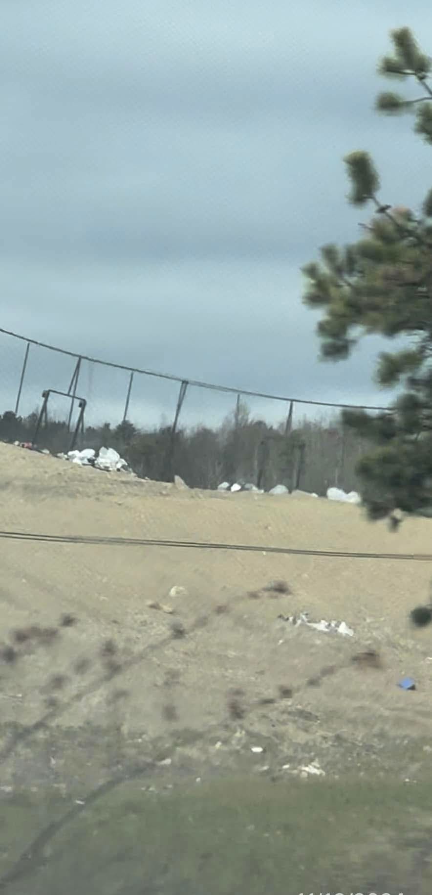 A chain-link perimeter fence with bags of trash piled against it on a barren landfill berm.