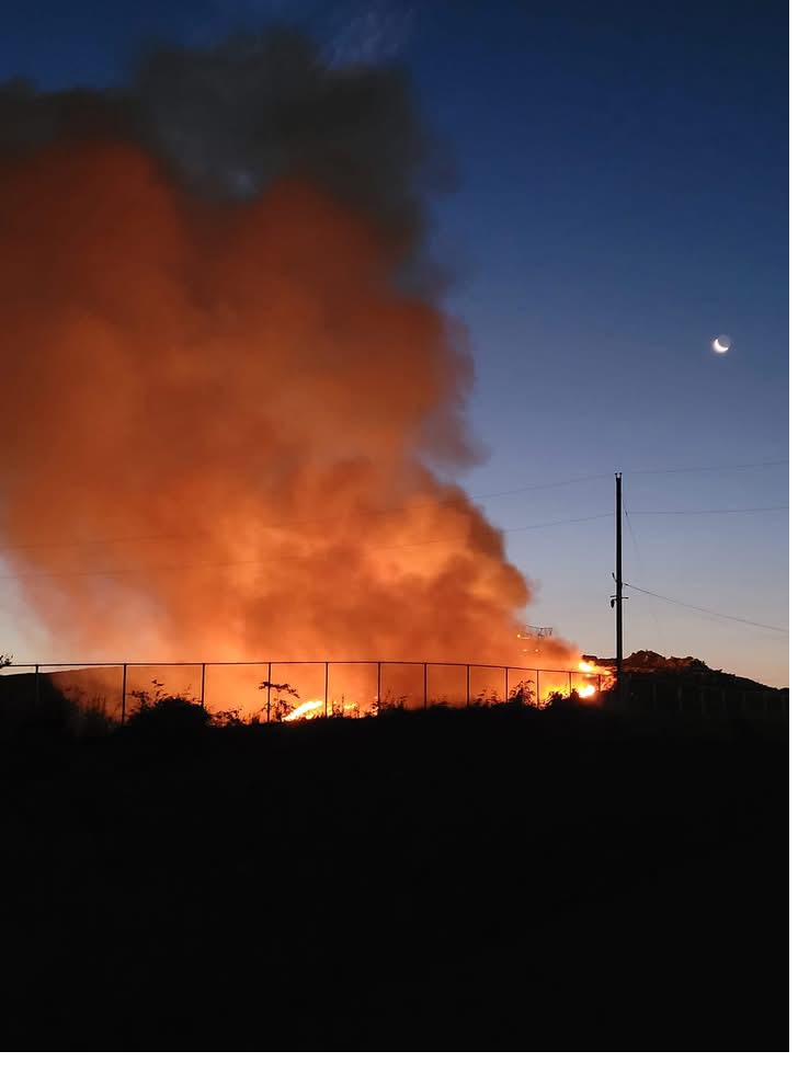 A landfill fire at dusk in Schuylkill County: an orange smoke plume rises into a deep navy sky above a perimeter fence.