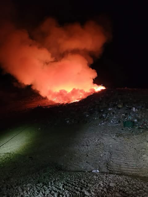A landfill fire at dusk: an orange plume rises into the deep blue sky over a perimeter fence.