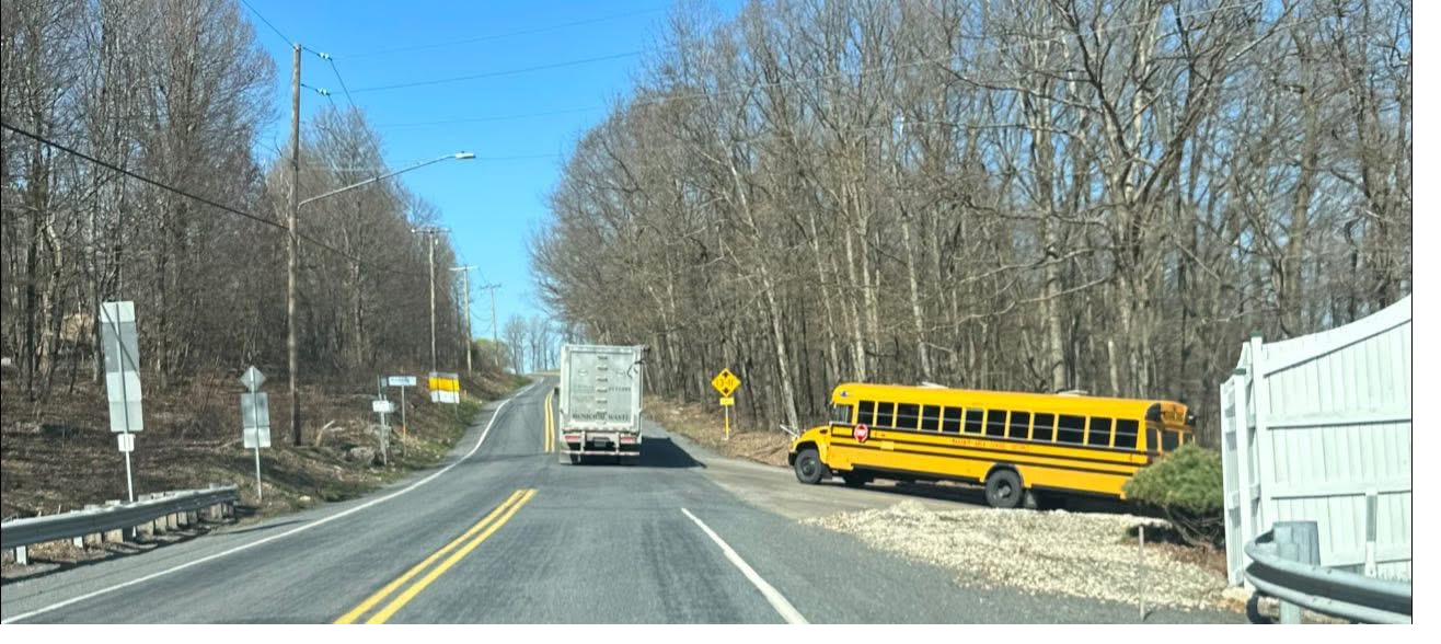 A rural road: a yellow school bus turning out of an industrial driveway as a municipal waste truck passes.
