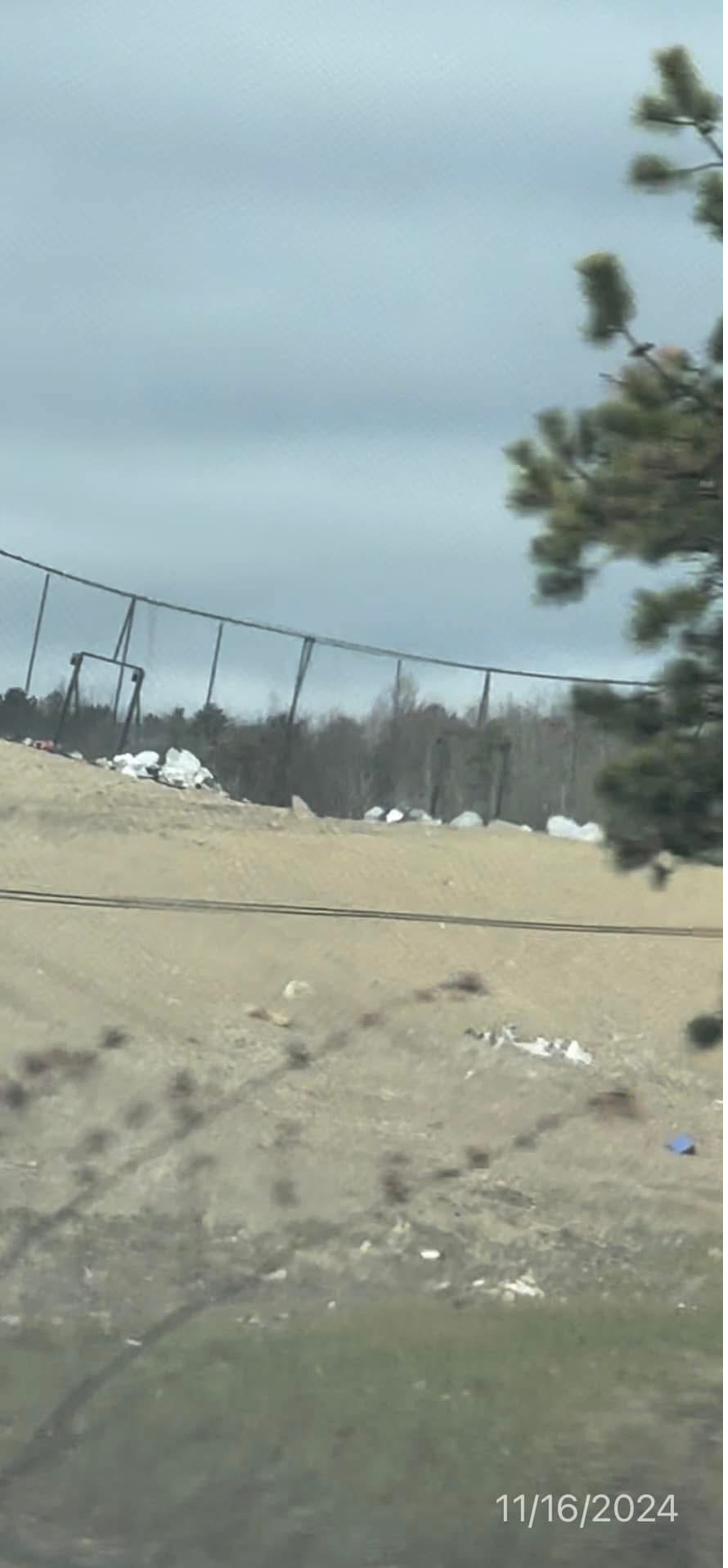 A landfill landscape: scattered debris along a perimeter fence with pine trees beyond.
